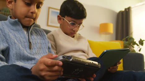 Dos-Niños-Sentados-En-Un-Sofá-En-Casa-Jugando-O-Transmitiendo-En-Una-Tableta-Digital-Y-Un-Dispositivo-De-Juegos-Portátil-1